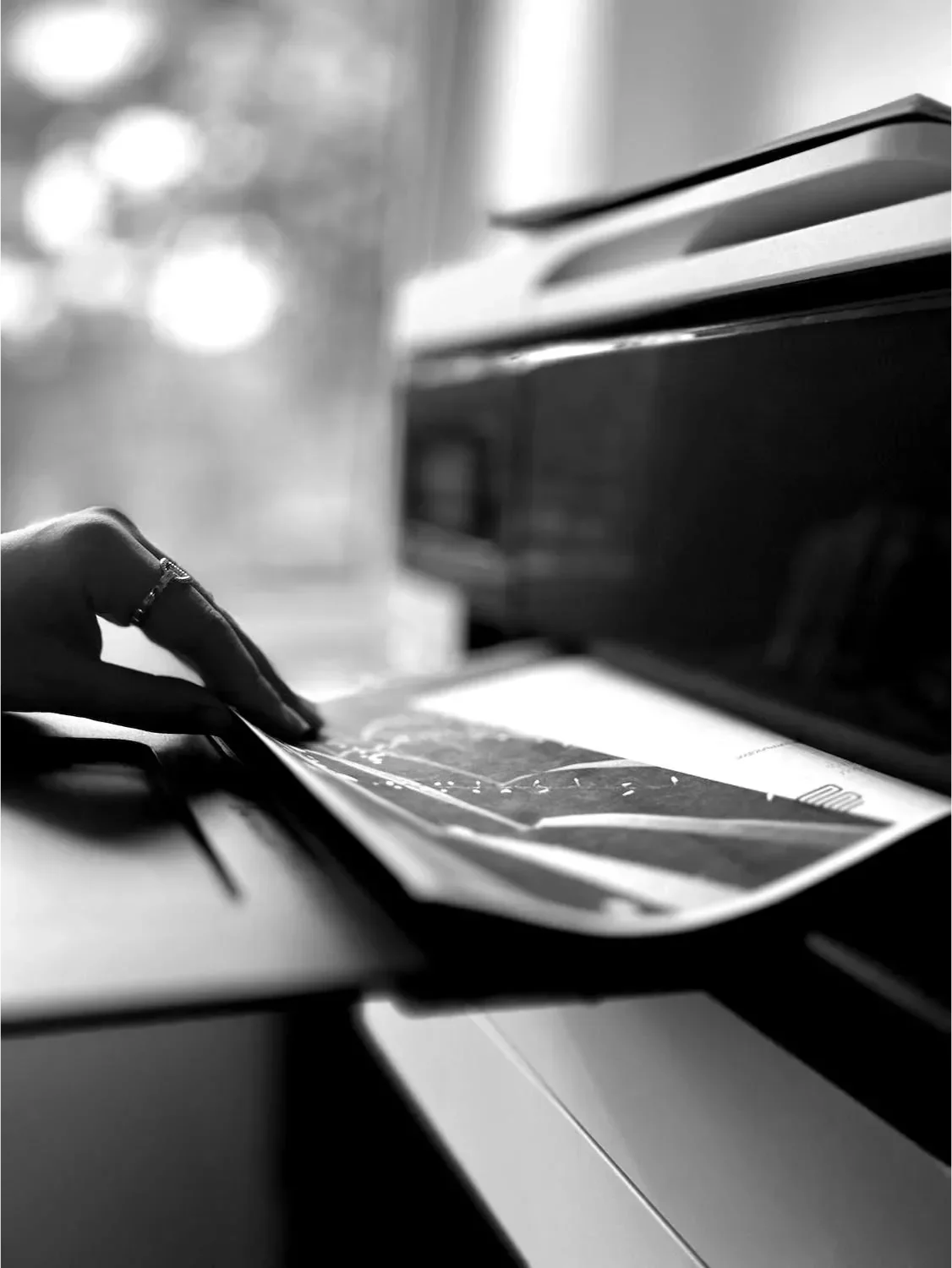 La main d’une personne place un document dans une imprimante ou un photocopieur, avec la machine en fonctionnement, une fenêtre en arrière-plan et un trombone noir posé à proximité.