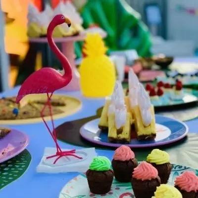 Une table avec des desserts assortis, dont des cupcakes et du cheesecake, décorée d'un ornement de flamant rose, d'une pièce maîtresse d'ananas jaune et d'un accent de trombone noir fantaisiste.