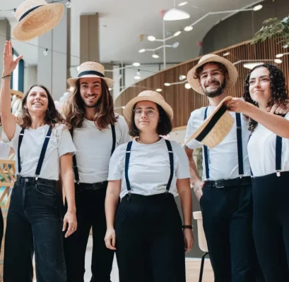 Un groupe de huit personnes portant des chemises blanches, des pantalons noirs, des bretelles et des chapeaux de paille se tiennent à l'intérieur lors d'un festival de Noël, certaines souriant et tenant leurs chapeaux.