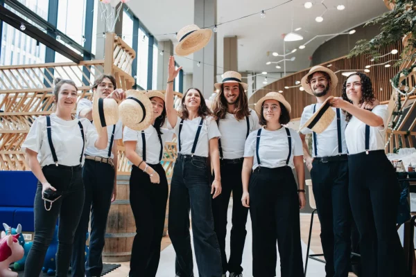 Un groupe de huit personnes portant des chemises blanches, des pantalons noirs, des bretelles et des chapeaux de paille se tiennent à l'intérieur lors d'un festival de Noël, certaines souriant et tenant leurs chapeaux.