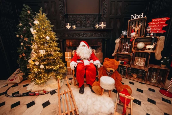 Le Père Noël est assis dans une salle festive près d'un arbre de Noël lumineux, entouré d'un gros ours en peluche, de cadeaux et d'un décor de vacances enchanteur, prêt à célébrer Noël.