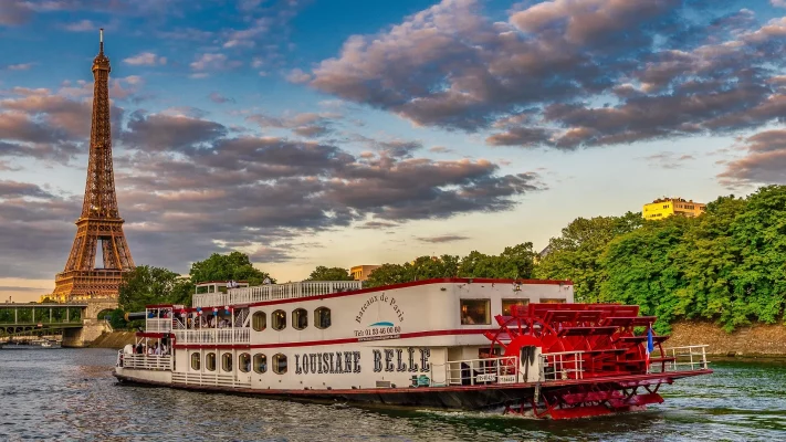 Un bateau à vapeur à aubes nommé Louisiane Belle propose une Croisière sur la Seine, glissant devant la Tour Eiffel et les arbres en arrière-plan sous un ciel partiellement nuageux.