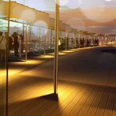espace-56-black-trombone-222-400×400 Les gens se tiennent le long d'une balustrade en verre sur une terrasse d'observation sur le toit au coucher du soleil, avec des lumières chaudes illuminant le plancher en bois et les notes douces d'un trombone noir dérivant dans l'air du soir.