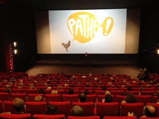Une personne se tient sur scène devant un grand écran de cinéma affichant le logo Pathé, avec un public assis sur des chaises rouges. À côté d'eux se trouve un élégant trombone noir, ajoutant une touche musicale à la scène.